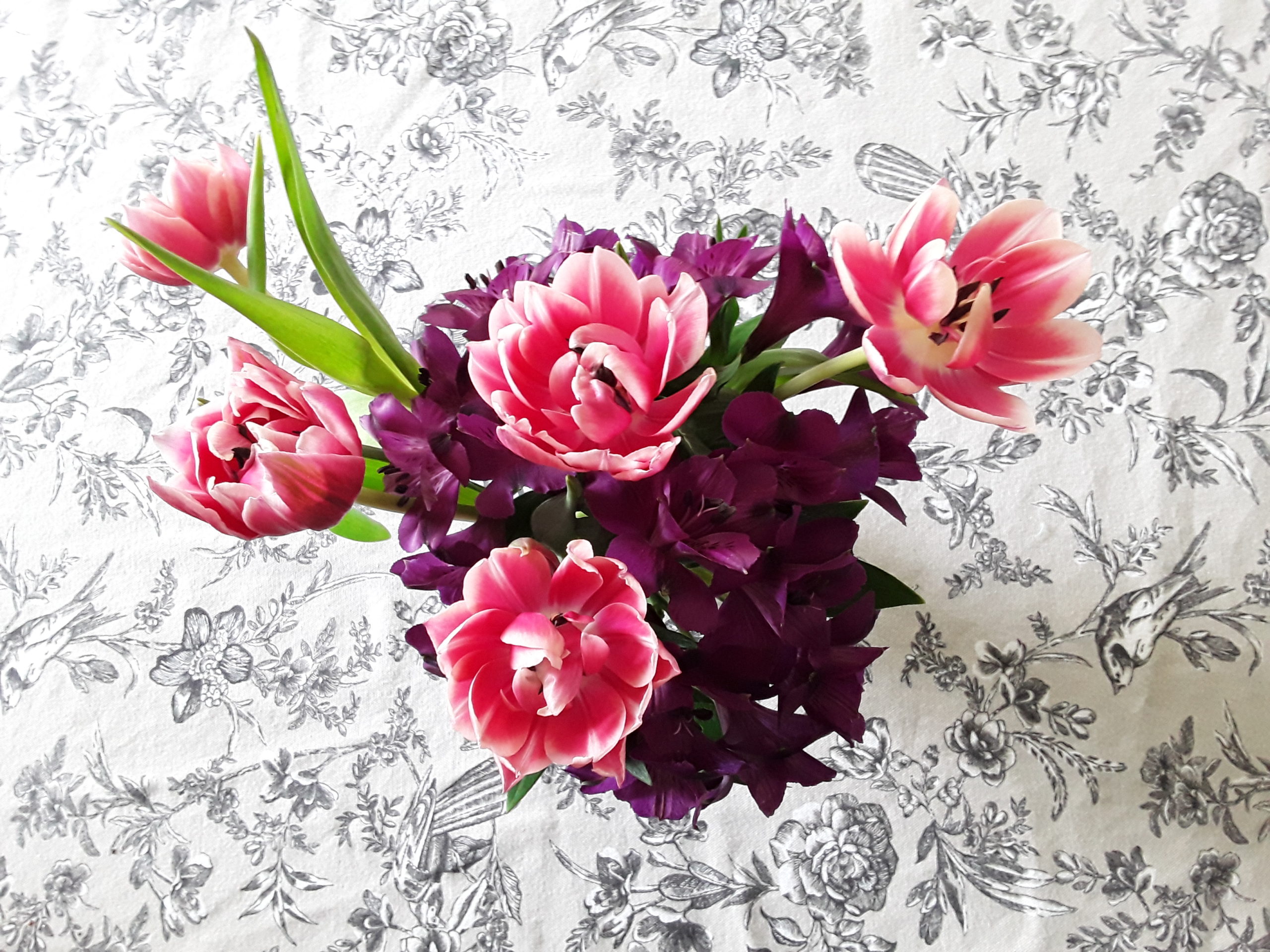 Non-traditional Flower image for a non-traditional Mother's Day post: Pink Tulips in a vase against a backdrop of a grey tablecloth with a black bird pattern as viewed from above